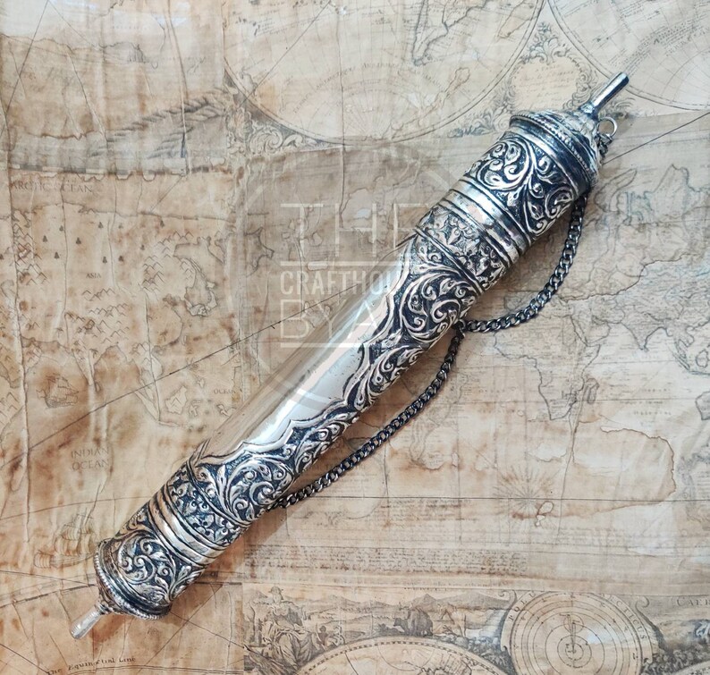 Pu&ograve; includere: Un astuccio argentato per pergamene con intricati motivi floreali e geometrici. La custodia cilindrica ha tappi decorativi e una catena. Lo sfondo &egrave; una mappa vintage.