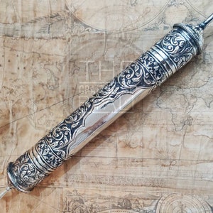 Pu&ograve; includere: Un astuccio per rotolo d'argento con incisioni floreali ornate. L'astuccio cilindrico ha tappi decorativi e una superficie lucida. Lo sfondo presenta una mappa vintage.