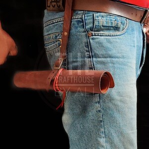 Op de afbeelding: Een bruin leren rolhoes met een rode band en gesp. Het hoesje is bevestigd aan een bruine leren riem met een metalen gesp. De riem wordt om de taille van een persoon gedragen die een blauwe jeans draagt.