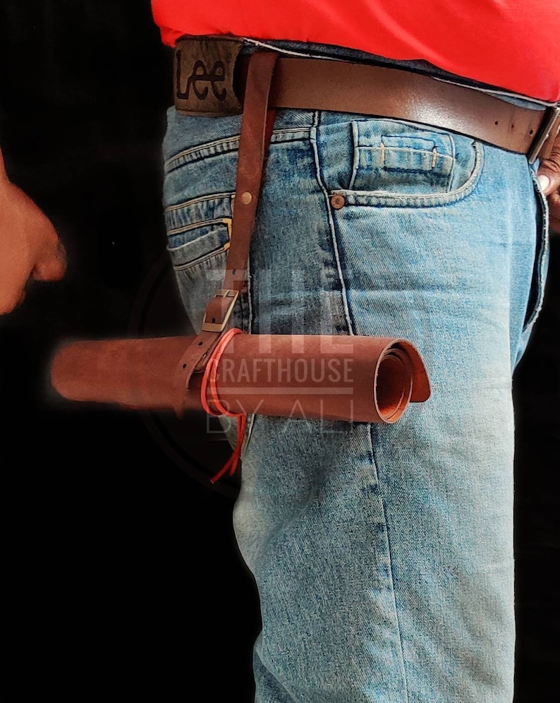 Op de afbeelding: Een bruin leren rolhoes met een rode band en gesp. Het hoesje is bevestigd aan een bruine leren riem met een metalen gesp. De riem wordt om de taille van een persoon gedragen die een blauwe jeans draagt.