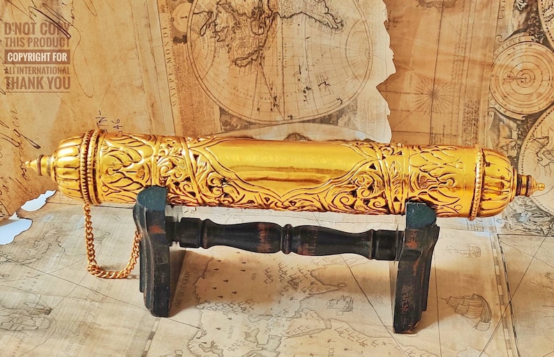 May include: A gold-colored, ornate, cylindrical container with intricate floral designs, resting on a dark wooden stand. The container has a chain attached to it.