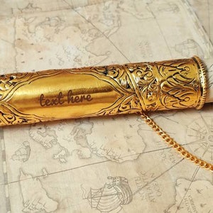May include: A gold metal scroll tube with intricate designs and a chain attached. The tube is open and a rolled piece of paper is inside. The text "text here" is engraved on the tube.