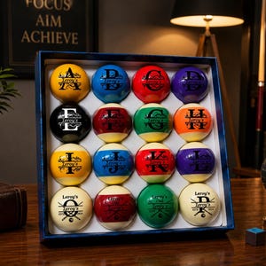 Op de afbeelding: Een set van zestien biljartballen gerangschikt in een vier-bij-vier raster in een blauwe displaydoos. Elke bal is voorzien van een unieke letter van A tot P, de tekst Leroy's en decoratieve gekruiste biljartkeus in zwarte inkt op het oppervlak.