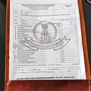 May include: A brown leather folder with a white paper document inside. The document is an Indian Income Tax Return Acknowledgement for the assessment year 2020-21. The document includes the taxpayer's name, address, PAN, and other details. The document also includes a stamp from the Income Tax Department.