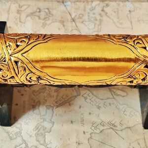 May include: A gold-colored metal scroll tube with intricate floral designs. The tube is on a wooden stand and has a small chain attached to the top.