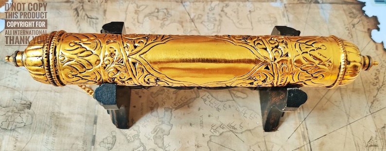 May include: A gold-colored metal scroll tube with intricate floral designs. The tube is on a wooden stand and has a small chain attached to the top.