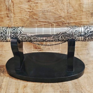 Pu&ograve; includere: Un astuccio argentato per rotoli con elaborati dettagli floreali, appoggiato su un supporto nero. L'astuccio cilindrico presenta estremit&agrave; decorative e una sezione centrale lucida. Una piccola catena &egrave; attaccata. Il supporto ha un design minimalista, lungo circa 30 cm.