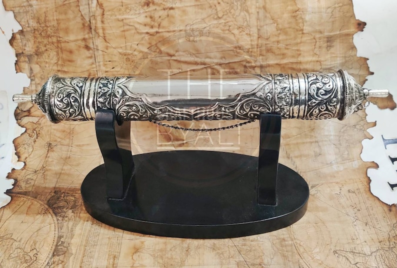 Pu&ograve; includere: Un astuccio argentato per rotoli con elaborati dettagli floreali, appoggiato su un supporto nero. L'astuccio cilindrico presenta estremit&agrave; decorative e una sezione centrale lucida. Una piccola catena &egrave; attaccata. Il supporto ha un design minimalista, lungo circa 30 cm.