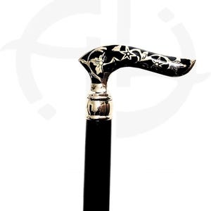 May include: A walking cane featuring a black shaft and a curved handle. The handle is black with an inlaid silver-colored floral pattern. A polished silver-colored metal collar connects the handle to the shaft, providing a smooth transition.
