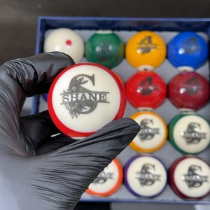 Op de afbeelding: Een biljartbal met een rode streep en een wit midden met een zwarte letter S en de naam SHANE in een schreeflettertype, geaccentueerd met florale lijntekeningen. De bal wordt voor een doos met andere kleurrijke biljartballen gehouden.