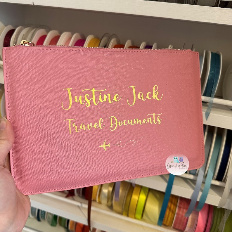 Pu&ograve; includere: Portadocumenti da viaggio rosa con cerniera dorata e il testo "Justine Jack Travel Documents" e due piccoli aerei con una linea tratteggiata tra loro. L'angolo in basso a destra ha un piccolo logo che dice "Georgina Ray Designs".