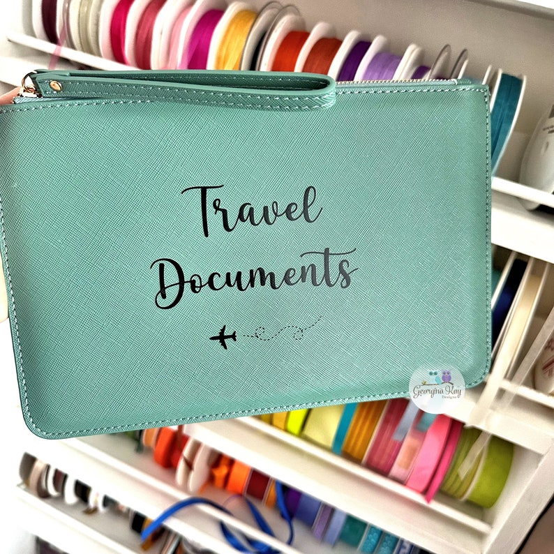 Pu&ograve; includere: Una pochette con cerniera color turchese con il testo "Travel Documents" e un piccolo grafico di un aereo. La pochette &egrave; realizzata in materiale testurizzato e ha una cerniera nera.
