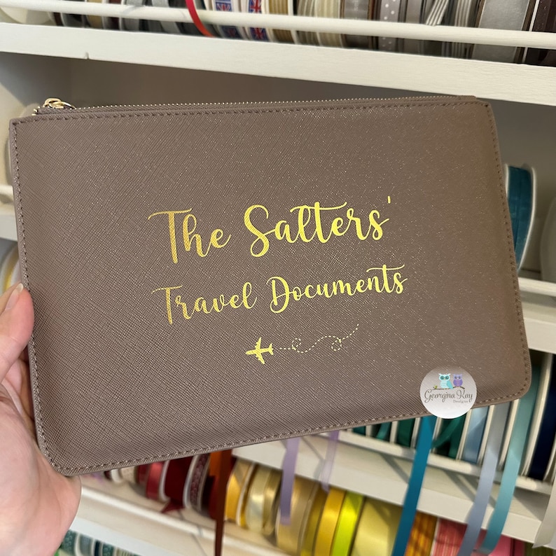 Pu&ograve; includere: Una pochette con cerniera marrone con lettere dorate che recitano "The Salters' Travel Documents" con una piccola grafica di aereo. La pochette ha un piccolo adesivo circolare bianco con il testo "Georgina Kay Designs" in lettere dorate.