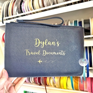 Pu&ograve; includere: Un portadocumenti da viaggio blu scuro con una cerniera dorata e una scritta dorata che recita "Dylan's Travel Documents" con un piccolo grafico di un aereo.