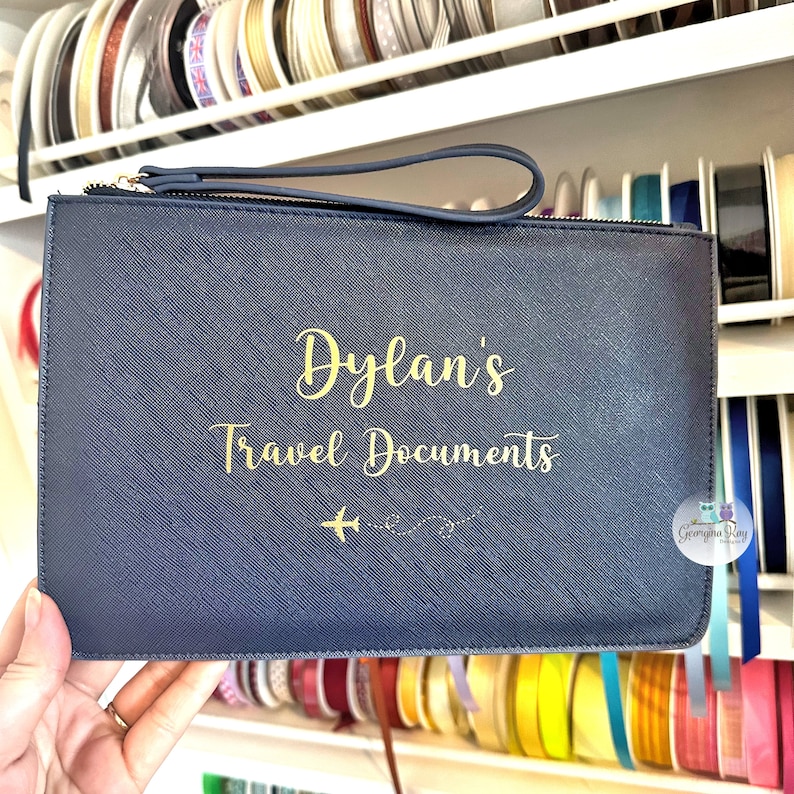 Pu&ograve; includere: Un portadocumenti da viaggio blu scuro con una cerniera dorata e una scritta dorata che recita "Dylan's Travel Documents" con un piccolo grafico di un aereo.