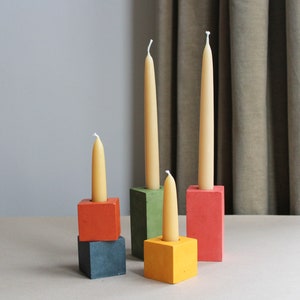 May include: A display of candles in various coloured holders. The holders are square or rectangular, in shades of red, green, yellow, and blue. The candles are cream-coloured, tall and slender. The arrangement is simple and elegant.