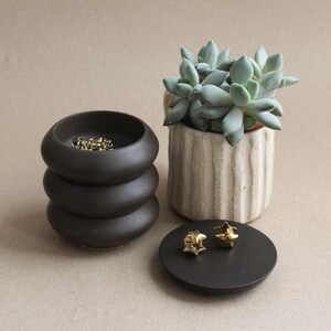 May include: A dark brown, three-tiered ceramic bowl containing small gold-toned chain links, a white ribbed ceramic planter holding a green succulent, and a flat, circular dark brown lid topped with two small, gold-toned star-shaped objects.