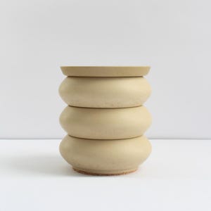 May include: A stackable ceramic container with a matte beige finish. The object consists of three rounded, bulbous sections stacked vertically, topped with a flat, circular lid. The surface has a subtle speckled texture throughout the exterior.