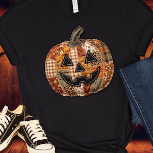 May include: A black short-sleeved t-shirt featuring a central graphic of a jack-o'-lantern pumpkin. The pumpkin is constructed from various fabric patches in shades of orange, brown, and tan, including plaid, floral, and leopard print patterns.