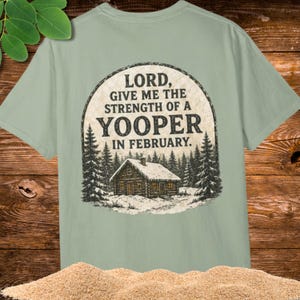 Puede incluir: Una camiseta verde salvia con un gráfico circular en la espalda. El diseño muestra una cabaña de madera nevada en un bosque con el texto: LORD, GIVE ME THE STRENGTH OF A YOOPER IN FEBRUARY. El estampado tiene una textura desgastada.