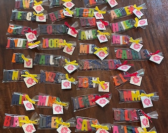 May include: A collection of colorful crayon name tags, each individually wrapped in clear plastic and tied with a yellow ribbon. The names include:  'Amy', 'Benjamin', 'Jamie', 'Pie', 'Lily', 'Drew', 'Beck', 'Do', 'Hale', 'Lora', 'Amy', 'Miles', 'Gabriel', 'Ryder', 'Jolie', 'Eli', 'Emma', 'Blake', 'Grey', 'Sally', 'Clark', 'Anna'.