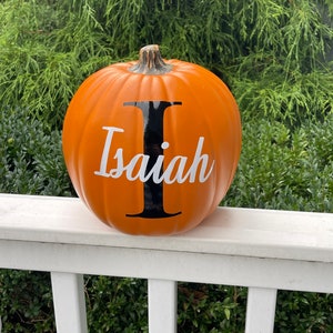 May include: A round orange artificial pumpkin featuring a large black letter I and the white script name Isaiah printed across the front. The pumpkin has a textured brown stem and a smooth matte finish, creating a distinct seasonal decoration.
