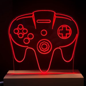 Op de afbeelding: Een rood neonlicht in de vorm van een klassieke videogamecontroller. De controller staat op een houten voet, tegen een zwarte achtergrond. Het ontwerp omvat knoppen, een D-pad en een centrale joystick.