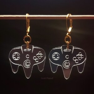 May include: A pair of gold-plated earrings featuring a clear acrylic design of a Nintendo 64 controller. The earrings are hanging from a gold hook.