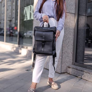 Puede incluir: Una mochila de cuero negro con cierre de solapa y correas ajustables. La mochila tiene un asa superior y un detalle de hebilla. La persona lleva una camisa a rayas azules y blancas, pantalones blancos y zapatos con estampado de serpiente.