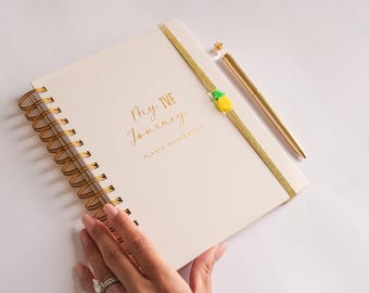 May include: A white spiral bound journal with a gold ribbon bookmark and a gold pineapple charm. The journal has the text "My IVF Journey" and "Flavia Wainwright" printed in gold on the cover.