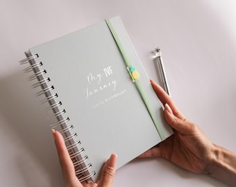 May include: A light blue spiral bound journal with the text "My IVF Journey" and the name "Olivia Wainwright" printed on the cover. A green ribbon is attached to the journal with a small pineapple charm.
