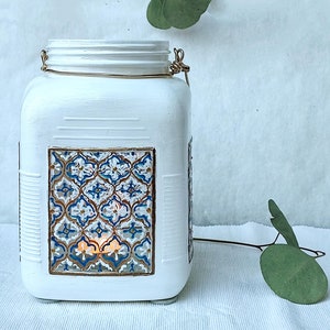 Może przedstawiać: Biały, malowany szklany słoik z uchwytem z złotego drutu i okienkiem z niebieskim i złotym wzorem. Słoik jest oświetlony od środka świeczką.