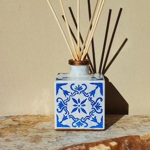 Op de afbeelding: Witte keramische diffuserfles met een blauw en gouden geometrisch patroon en houten diffuserstokjes.