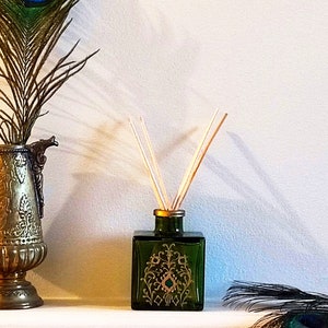 May include: A green glass diffuser bottle with gold accents and a decorative design. The bottle has wooden sticks inserted into the top and is sitting on a white surface. A peacock feather is in the background.