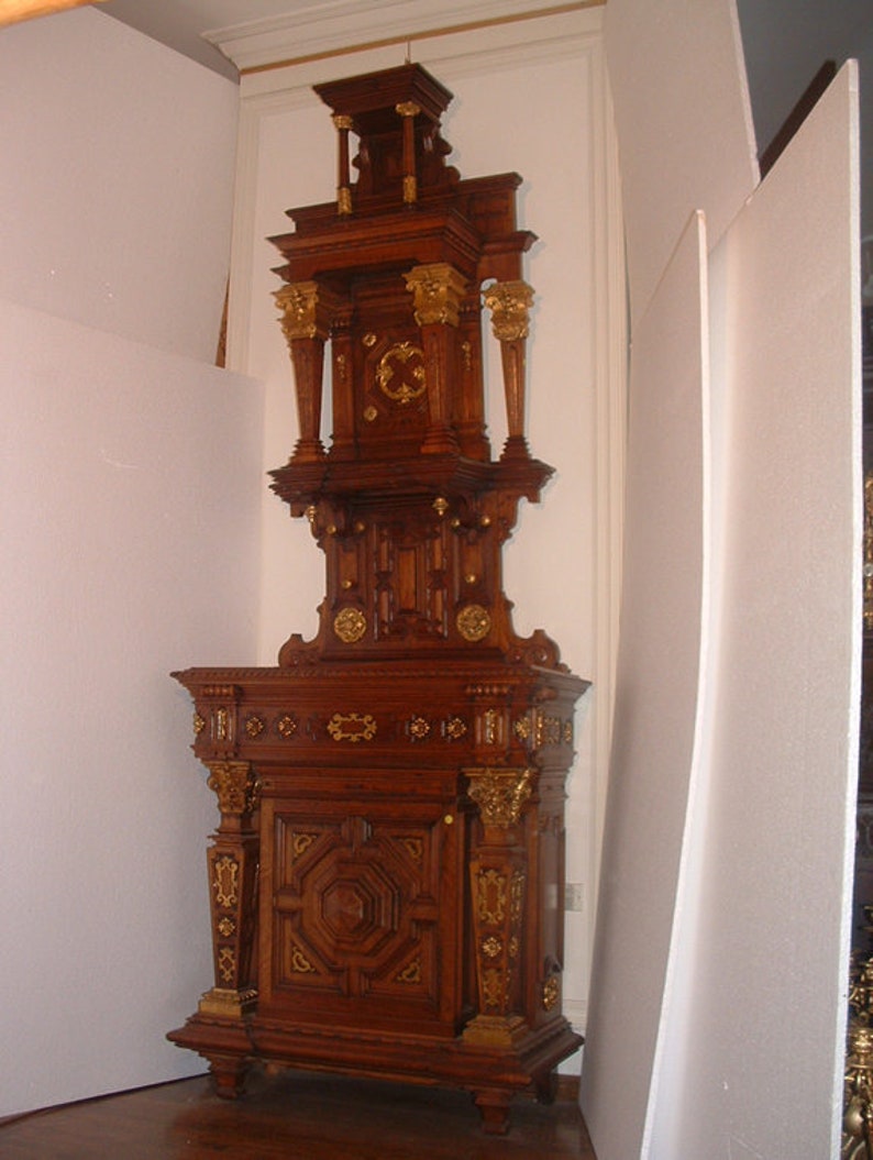 May include: A tall, ornately carved wooden cabinet with gold accents. The cabinet has a decorative top with a central design and two side columns. The lower section has a single door with a geometric pattern and is flanked by two decorative columns.