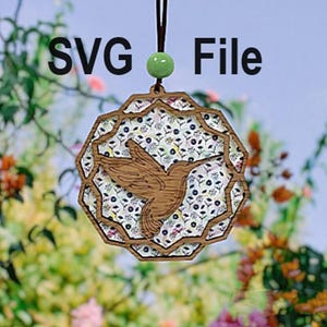 May include: A wooden hummingbird ornament with a floral pattern, hanging from a brown cord with a green bead. The ornament is shaped like a circle with a hummingbird cut out in the center. The hummingbird is facing right and has its wings spread out. The floral pattern is in shades of blue, pink, and green.