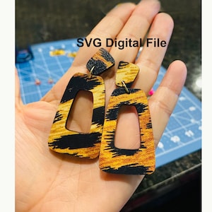 May include: A pair of geometric wooden earrings with a black and gold animal print design. The earrings have a cutout shape and are hanging from a person's hand. SVG Digital File.