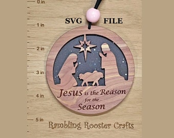 May include: Circular wooden Christmas ornament with a nativity scene design. The ornament features a star, figures, and the text "Jesus is the Reason for the Season." It is approximately 10 cm in diameter and has a black cord and pink bead for hanging.