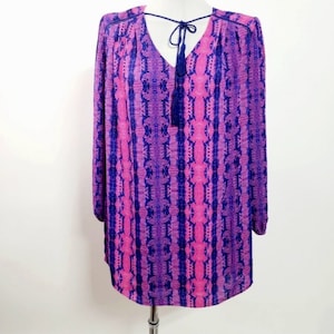 May include: A pink and purple patterned blouse with a blue tassel tie at the neckline. The blouse has long sleeves and a relaxed fit.