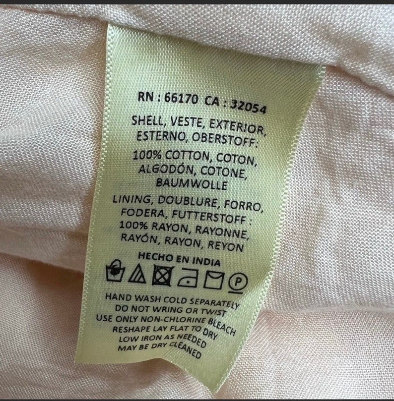 May include: A beige garment care label with the text "RN: 66170 CA: 32054" and care instructions for hand washing cold separately, do not wring or twist, use only non-chlorine bleach, reshape lay flat to dry, low iron as needed, may be dry cleaned. The label also includes the text "HECHO EN INDIA".