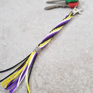 K&ouml;nnte beinhalten: Ein geflochtener Schl&uuml;sselanh&auml;nger in Lila, Wei&szlig; und Gelb mit einer silbernen Perle und einem Quasten. Der Schl&uuml;sselanh&auml;nger ist an einem Schl&uuml;sselsatz mit gr&uuml;nen und roten Schl&uuml;sselkappen befestigt.