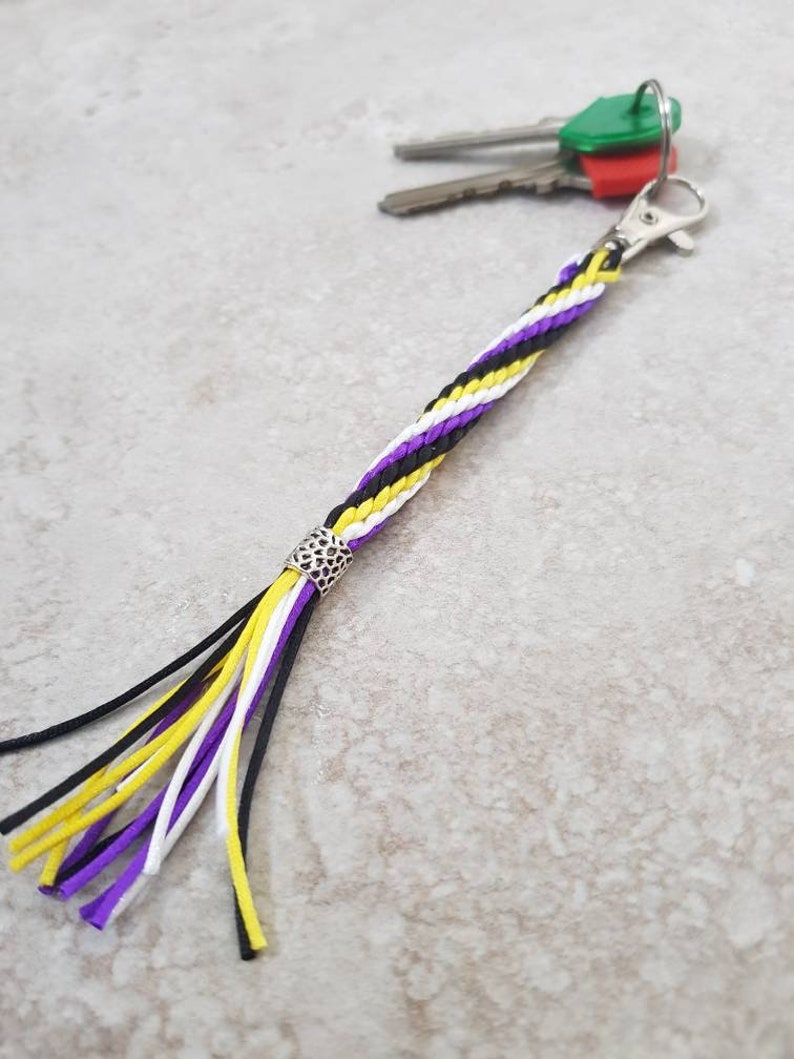 K&ouml;nnte beinhalten: Ein geflochtener Schl&uuml;sselanh&auml;nger in Lila, Wei&szlig; und Gelb mit einer silbernen Perle und einem Quasten. Der Schl&uuml;sselanh&auml;nger ist an einem Schl&uuml;sselsatz mit gr&uuml;nen und roten Schl&uuml;sselkappen befestigt.