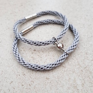 May include: Two grey braided cord bracelets with silver magnetic clasps. One bracelet has a silver charm with a small ball attached.