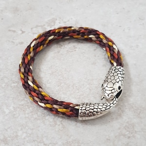 May include: Braided bracelet with a snake head clasp. The bracelet features a mix of brown, yellow, orange, and cream-coloured threads. The snake head clasp is silver-coloured and detailed.