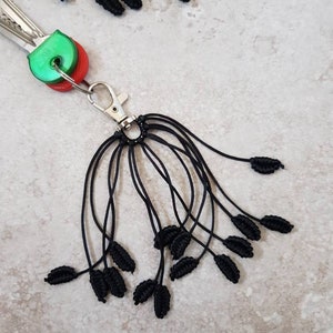May include: A black macrame keychain with a silver-colored clasp, holding keys with green and red accents. The keychain features multiple black cord strands, each ending in a leaf-shaped macrame knot. The background is a light, speckled surface.