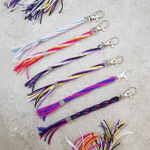 K&ouml;nnte beinhalten: Ein Set aus sieben bunten geflochtenen Schl&uuml;sselanh&auml;ngern mit silbernen Schl&uuml;sselringen und Quasten. Jeder Schl&uuml;sselanh&auml;nger hat eine andere Farbkombination, darunter Lila, Wei&szlig;, Gelb, Orange, Rosa, Blau und Schwarz.