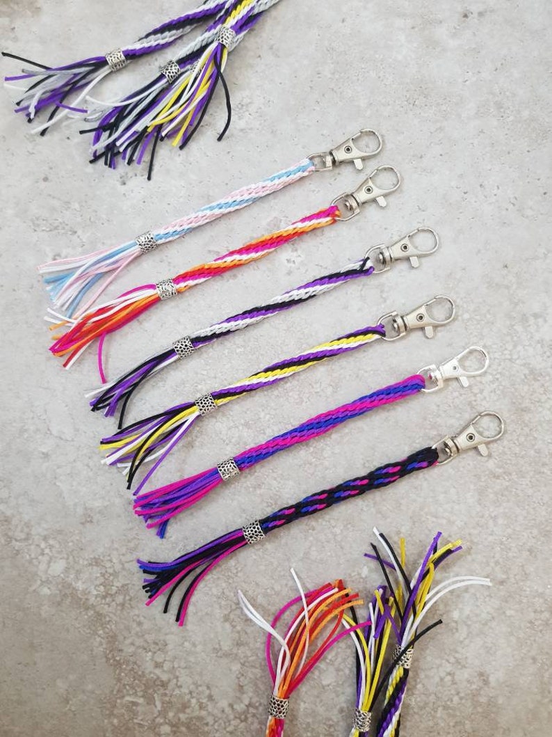 K&ouml;nnte beinhalten: Ein Set aus sieben bunten geflochtenen Schl&uuml;sselanh&auml;ngern mit silbernen Schl&uuml;sselringen und Quasten. Jeder Schl&uuml;sselanh&auml;nger hat eine andere Farbkombination, darunter Lila, Wei&szlig;, Gelb, Orange, Rosa, Blau und Schwarz.