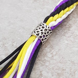 K&ouml;nnte beinhalten: Eine mehrfarbige geflochtene Schnur mit einer silbernen Metallperle. Die Schnur ist schwarz, wei&szlig;, gelb und lila.