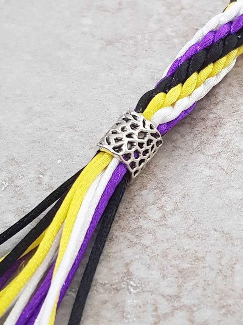 K&ouml;nnte beinhalten: Eine mehrfarbige geflochtene Schnur mit einer silbernen Metallperle. Die Schnur ist schwarz, wei&szlig;, gelb und lila.