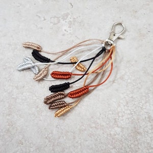 May include: A silver key chain with multiple strands of braided cord in brown, beige, gold, and black colours. The cords are tied in small knots at the ends.